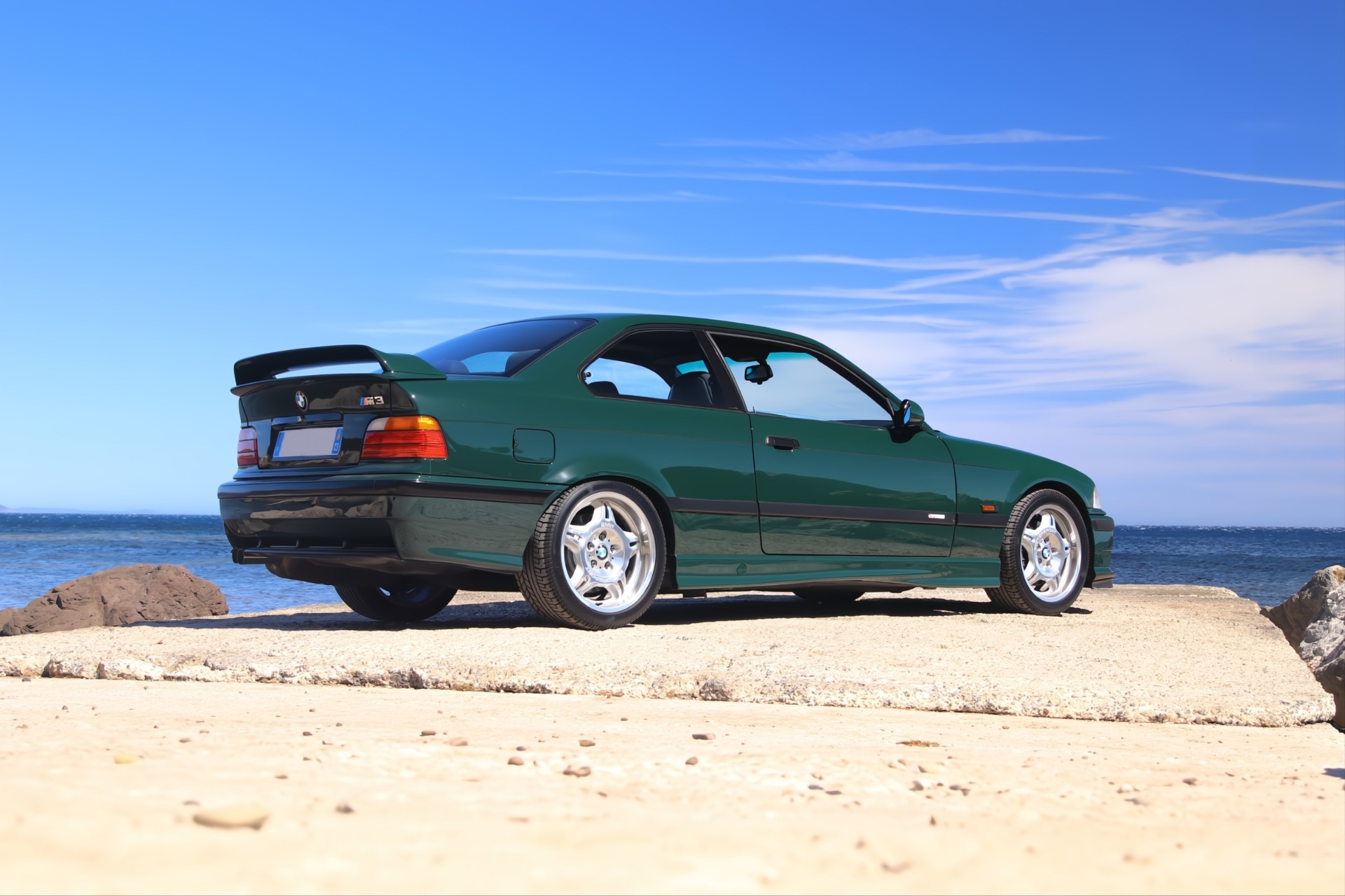 BMW M3 GT E36 de trois-quarts arrière face à la mer, ciel bleu