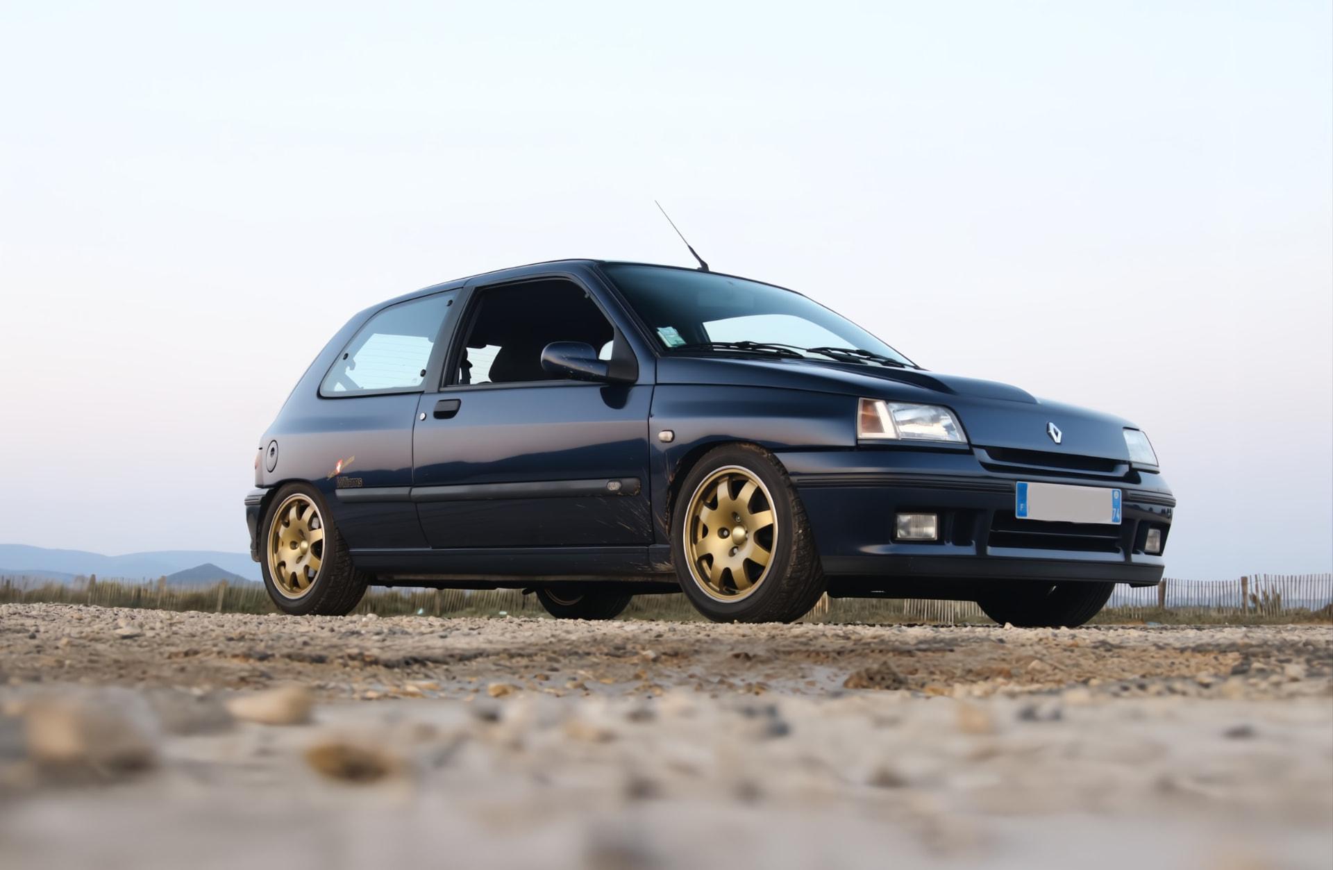 Renault Clio Williams bleu profond aux jantes dorées, garée en bord d'étang au coucher du soleil