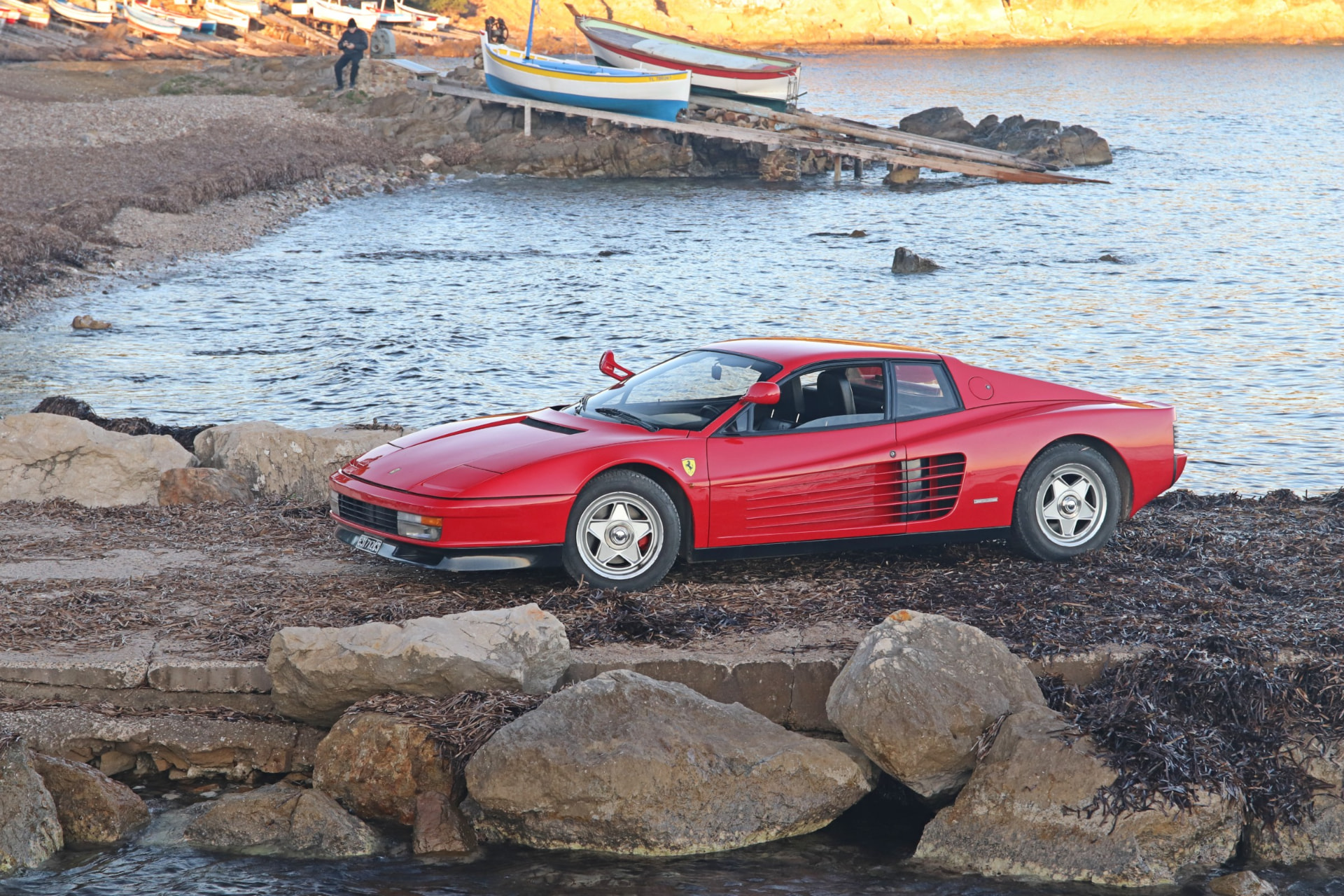 Ferrari Testarossa monospecchio rouge de profil devant des barques de pêche